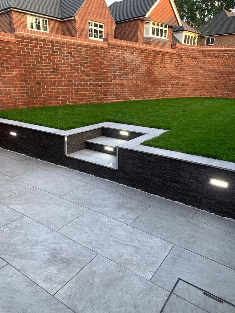 Grey porcelain patio with steps up to a raised lawn behind a dark retaining wall with built-in lights